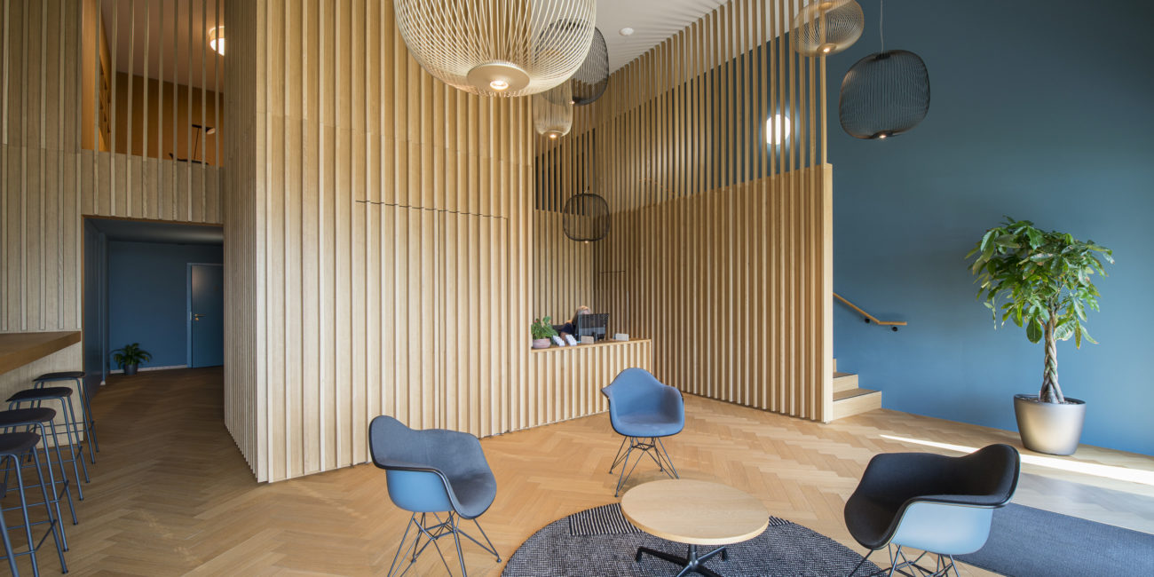 Un hall d'entrée moderne orné de lattes en bois massif aux murs, de chaises bleues disposées autour d'un tapis circulaire avec des tables basses, d'un coin bar avec des tabourets, de grandes suspensions lumineuses et d'une plante en pot près de l'escalier, pour un agencement intérieur élégant. - Menuiserie Ledermann SA
