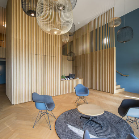 Un hall d'entrée moderne orné de lattes en bois massif aux murs, de chaises bleues disposées autour d'un tapis circulaire avec des tables basses, d'un coin bar avec des tabourets, de grandes suspensions lumineuses et d'une plante en pot près de l'escalier, pour un agencement intérieur élégant. - Menuiserie Ledermann SA