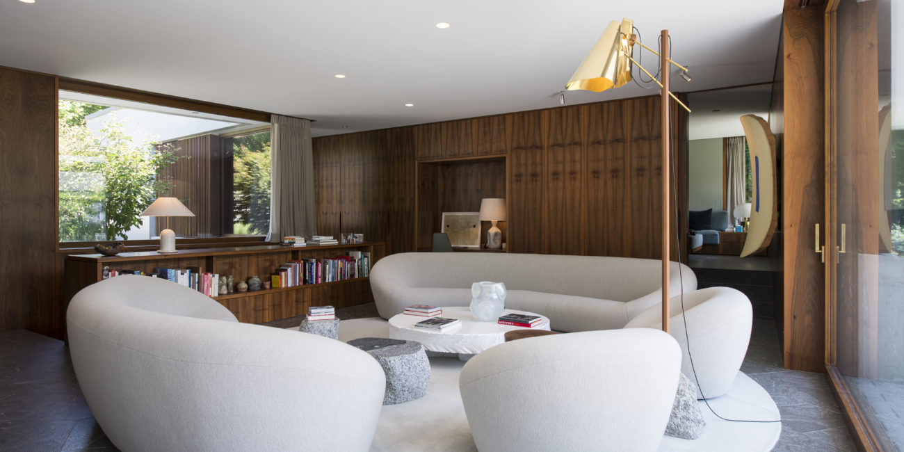Un salon moderne avec des canapés gris clair aux lignes arrondies disposés autour d'une table basse ronde blanche, le tout posé sur un tapis circulaire. L'espace se caractérise par des murs lambrissés en bois massif, des bibliothèques encastrées et de grandes fenêtres laissant entrer la lumière naturelle. - Menuiserie Ledermann SA