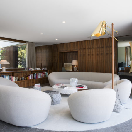 Un salon moderne avec des canapés gris clair aux lignes arrondies disposés autour d'une table basse ronde blanche, le tout posé sur un tapis circulaire. L'espace se caractérise par des murs lambrissés en bois massif, des bibliothèques encastrées et de grandes fenêtres laissant entrer la lumière naturelle. - Menuiserie Ledermann SA