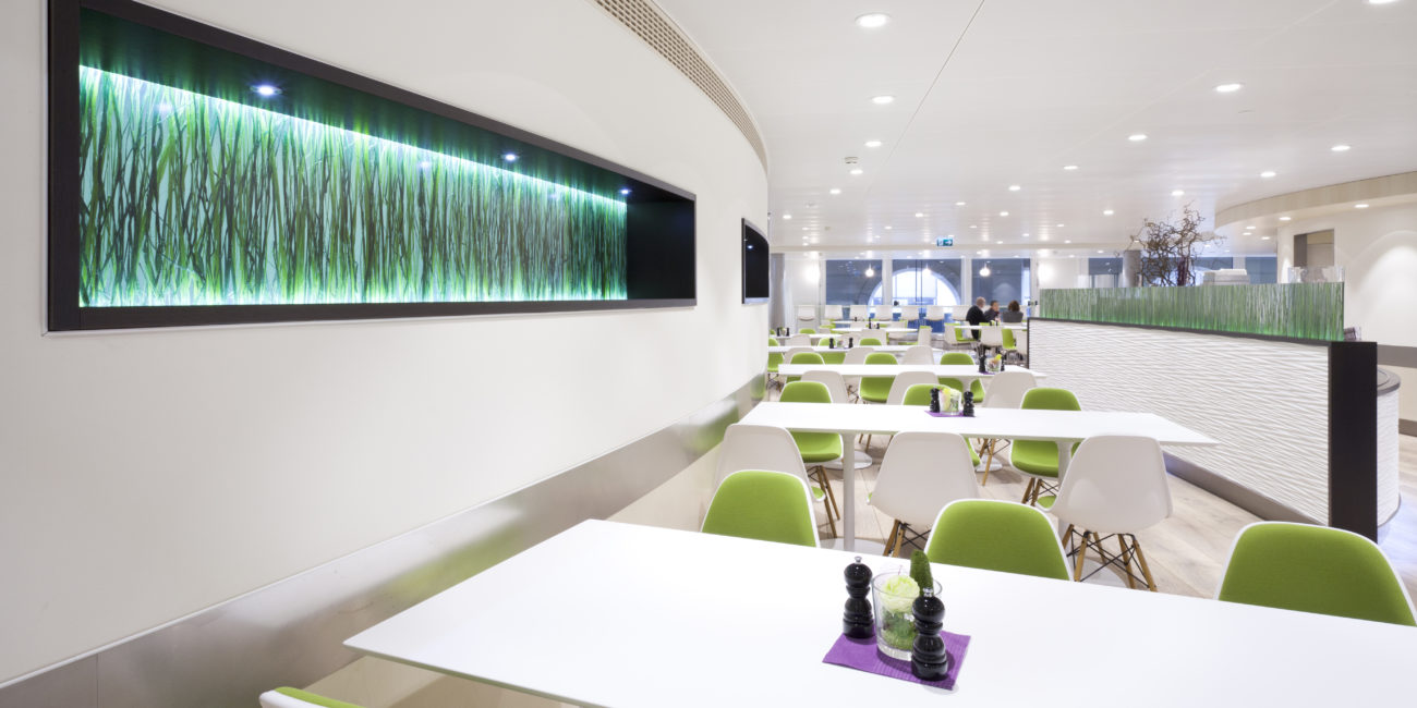 Une cafétéria moderne dotée de tables blanches et de chaises vertes, agrémentée de touches de bois massif et d'une décoration murale composée de motifs verts lumineux évoquant de l'herbe. Cet espace lumineux et épuré propose un dressage de table minimaliste, avec des salières et poivrières. - Menuiserie Ledermann SA