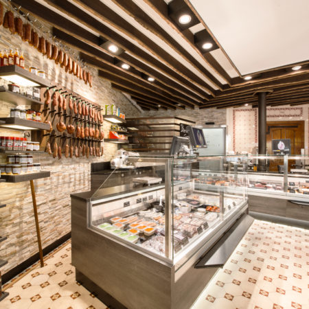 Intérieur moderne d'une épicerie fine, caractérisé par un agencement comprenant un comptoir vitré présentant un assortiment de charcuteries, des étagères sur lesquelles sont suspendues des saucisses séchées, ainsi que des touches de bois massif qui contrastent avec les murs en pierre et le sol en carrelage à motifs. - Menuiserie Ledermann SA