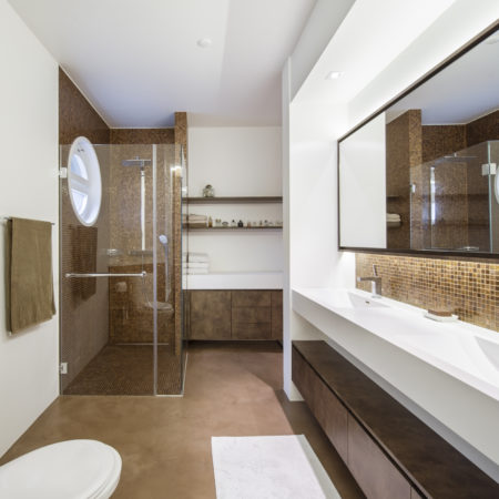 Salle de bains moderne avec un carrelage en mosaïque marron dans l'espace douche, une porte de douche en verre, une double vasque avec de grands miroirs, des meubles suspendus en bois massif, des toilettes blanches et une fenêtre ronde. Les murs et les étagères blancs accueillent les serviettes et les articles de toilette. - Menuiserie Ledermann SA