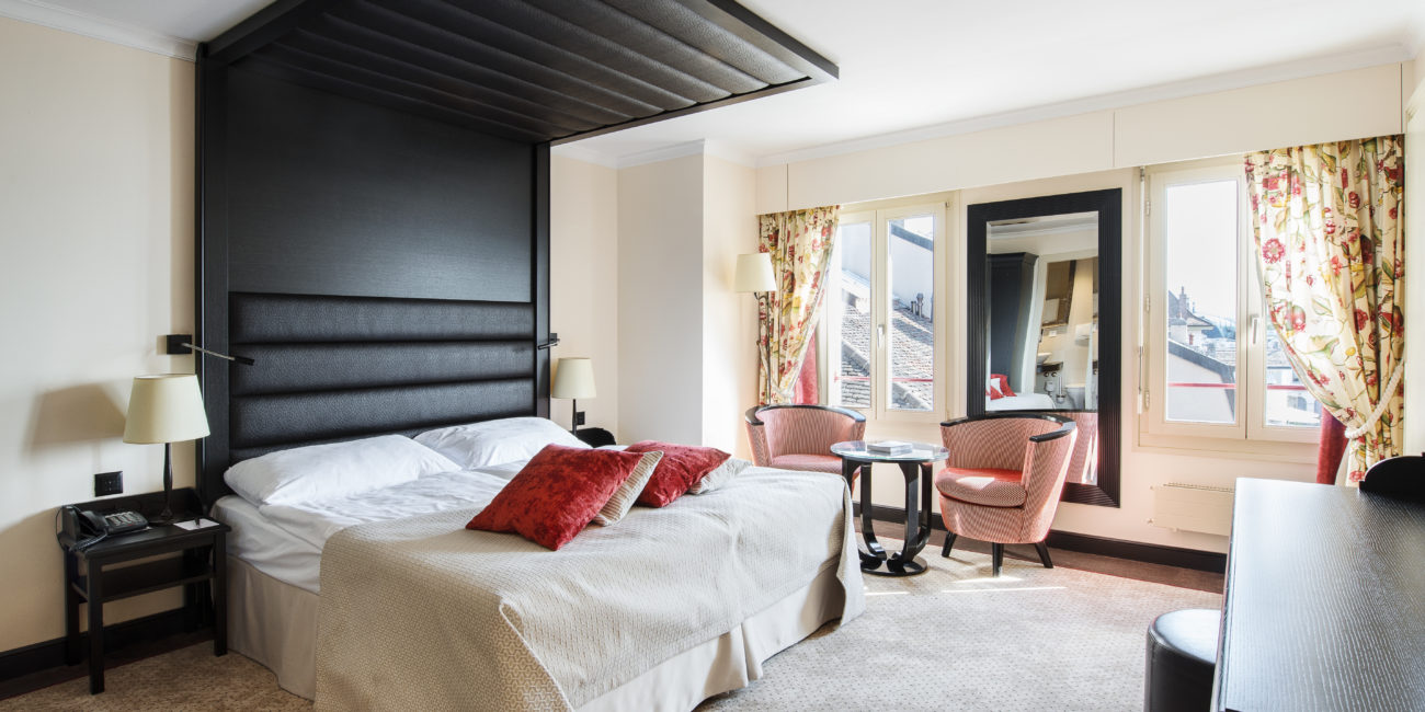 Une chambre d'hôtel avec un grand lit, deux oreillers rouges, une tête de lit noire, un agencement intérieur sur mesure comprenant une petite table ronde avec deux chaises, un bureau, un miroir, des fenêtres ornées de rideaux à motifs floraux et de la moquette beige. - Menuiserie Ledermann SA