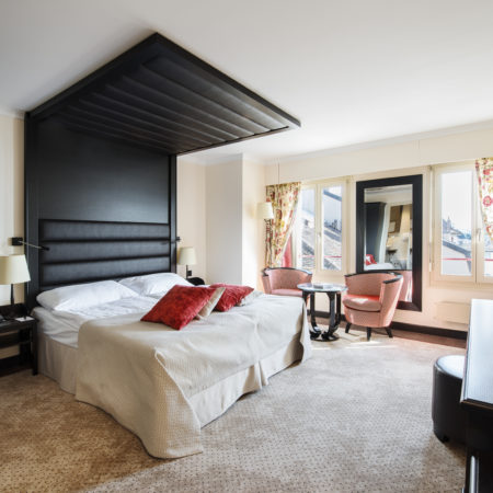 Une chambre d'hôtel avec un grand lit, deux oreillers rouges, une tête de lit noire, un agencement intérieur sur mesure comprenant une petite table ronde avec deux chaises, un bureau, un miroir, des fenêtres ornées de rideaux à motifs floraux et de la moquette beige. - Menuiserie Ledermann SA