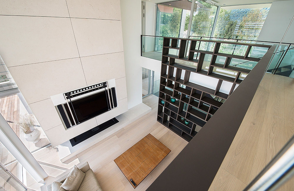 Un salon moderne sur deux niveaux, doté d'une étagère en menuiserie sur mesure, d'une télévision fixée au mur au-dessus d'une cheminée, d'un parquet en bois massif clair, d'une table basse rectangulaire et de grandes fenêtres offrant une vue sur l'extérieur. - Menuiserie Ledermann SA