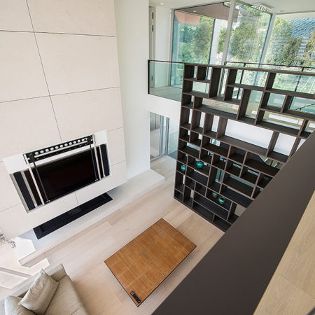 Un salon moderne sur deux niveaux, doté d'une étagère en menuiserie sur mesure, d'une télévision fixée au mur au-dessus d'une cheminée, d'un parquet en bois massif clair, d'une table basse rectangulaire et de grandes fenêtres offrant une vue sur l'extérieur. - Menuiserie Ledermann SA