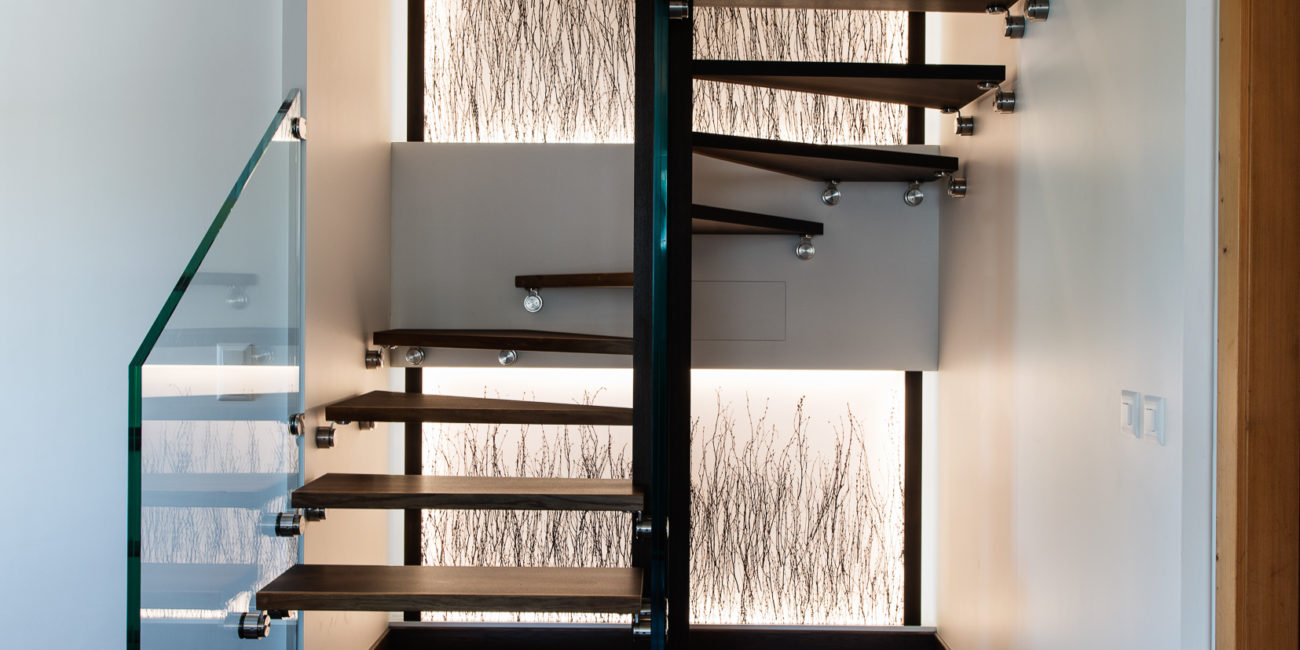 Escalier flottant moderne composé de marches en bois massif foncé, d'une rampe en verre sur la gauche et d'un mur décoratif orné de lignes verticales abstraites, s'intégrant dans un agencement intérieur contemporain avec un parquet en bois foncé assorti. - Menuiserie Ledermann SA