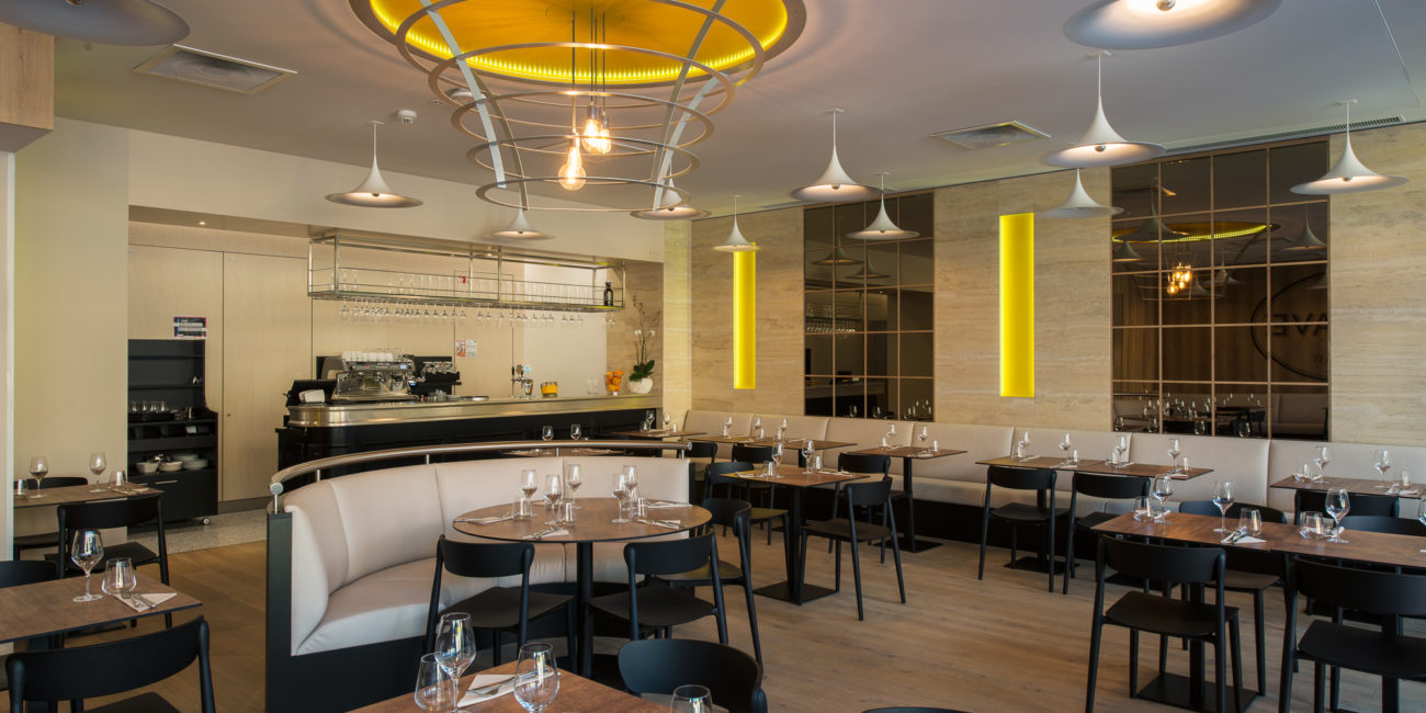 L'intérieur d'un restaurant moderne se caractérise par des tables dressées avec de la verrerie et des couverts, des chaises noires, des banquettes beiges, un parquet en bois massif et des luminaires contemporains aux reflets jaunes. L'espace bar est visible à l'arrière-plan. - Menuiserie Ledermann SA