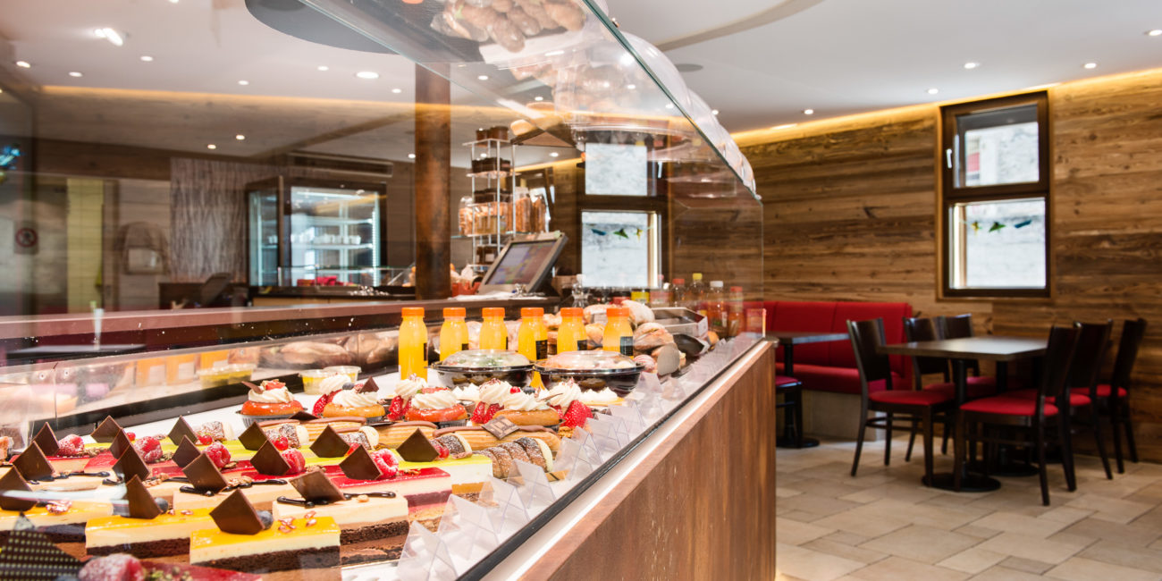 Une vitrine en verre dans une boulangerie ou un café présente un assortiment de gâteaux, de pâtisseries et de desserts. Des boissons en bouteille sont alignées derrière la vitrine. Des chaises et des tables rouges sont disposées dans un coin salon aux lumières vives, aux murs en bois massif, créant ainsi un agencement intérieur chaleureux. - Menuiserie Ledermann SA