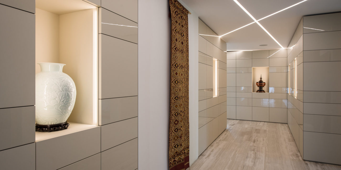 Un couloir moderne aux murs carrelés brillants, avec des étagères encastrées sur lesquelles sont disposés des vases décoratifs, un tapis à motifs accroché au mur et un parquet en bois massif clair. Les plafonniers forment des lignes qui se croisent au-dessus, créant ainsi un agencement intérieur élégant. - Menuiserie Ledermann SA