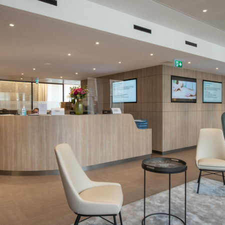 Un espace d'accueil moderne comprenant des boiseries en bois massif, deux fauteuils couleur crème, une petite table ronde, un fauteuil bleu, des écrans numériques fixés au mur, ainsi qu'un comptoir d'accueil orné de fleurs et de panneaux d'information. - Menuiserie Ledermann SA