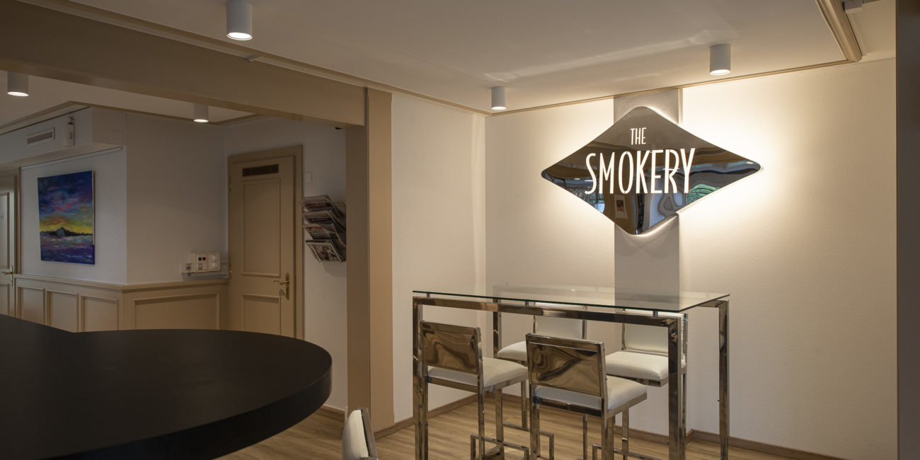 Un coin salon moderne avec une table en verre et trois chaises, sous une enseigne murale portant l'inscription « THE SMOKERY ». La pièce se distingue par son parquet en bois massif, ses murs beiges et son éclairage discret au plafond, qui mettent en valeur un agencement intérieur raffiné. - Menuiserie Ledermann SA