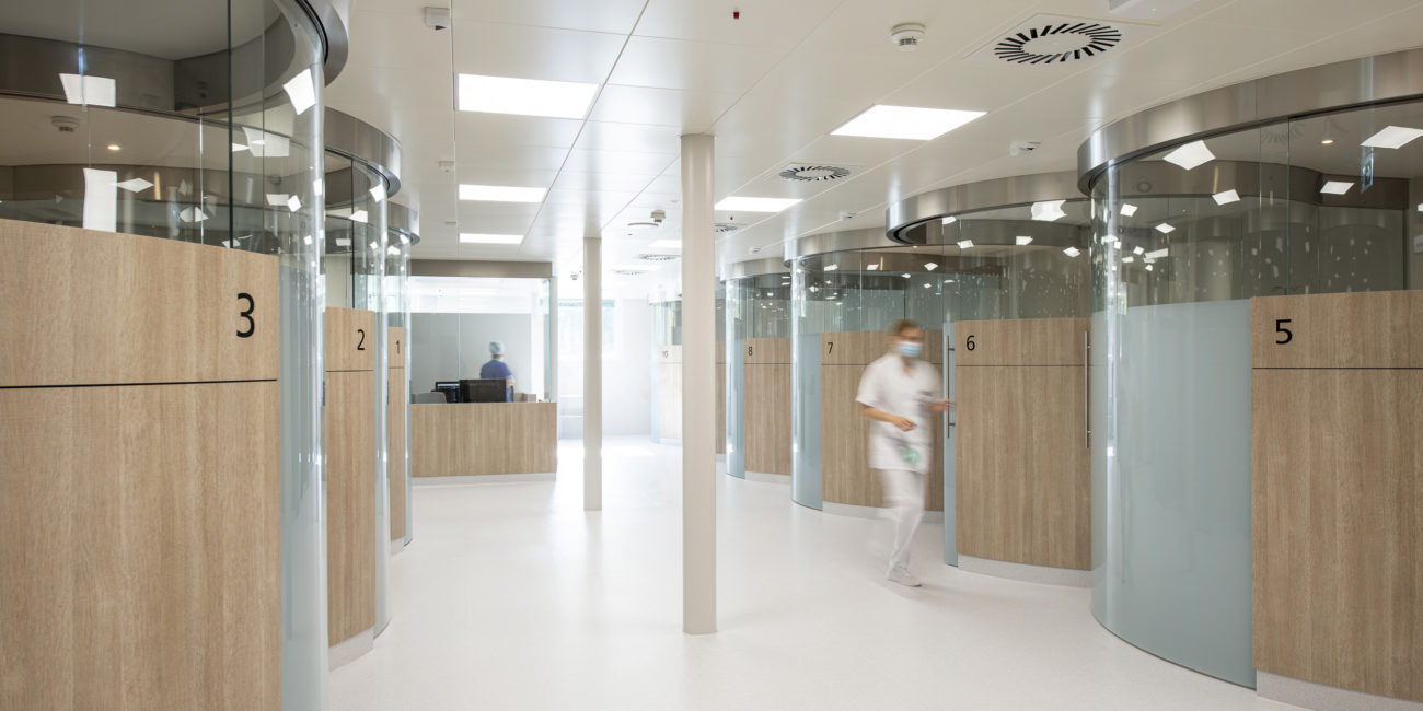 L'intérieur d'un hôpital moderne se caractérise par des chambres de patients numérotées aux parois vitrées et d'élégants lambris en bois massif, qui mettent en valeur un travail de menuiserie sur mesure d'une grande qualité. Un membre du personnel soignant en blouse blanche passe devant une autre personne qui travaille à un bureau dans cet espace propre et baigné de lumière. - Menuiserie Ledermann SA