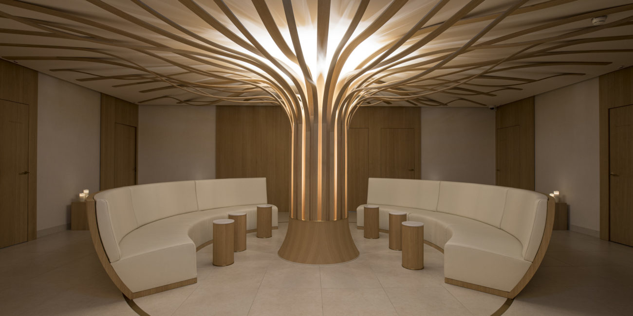 Un salon moderne avec des canapés beiges aux lignes courbes disposés en demi-cercle autour d'un pilier sculptural en bois massif évoquant un arbre, agrémenté d'éléments en bois ramifiés qui s'étendent jusqu'au plafond ; de petites tables cylindriques viennent rehausser cet agencement intérieur raffiné. - Menuiserie Ledermann SA