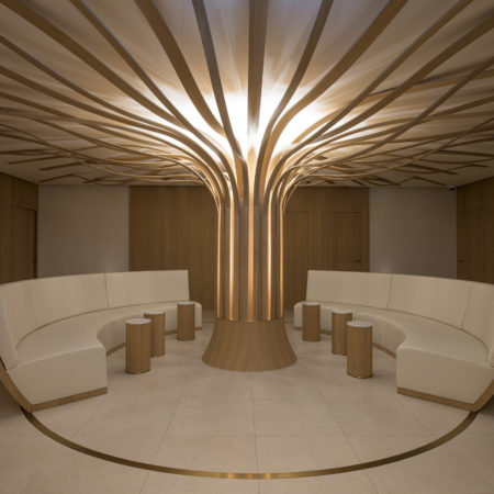 Un salon moderne avec des canapés beiges aux lignes courbes disposés en demi-cercle autour d'un pilier sculptural en bois massif évoquant un arbre, agrémenté d'éléments en bois ramifiés qui s'étendent jusqu'au plafond ; de petites tables cylindriques viennent rehausser cet agencement intérieur raffiné. - Menuiserie Ledermann SA