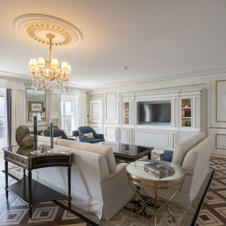 Un salon élégant avec un parquet en bois massif à motifs, des canapés couleur crème, une table noire, une table basse en verre, une télévision murale, un lustre et de grandes fenêtres donnant sur les arbres et l'eau, pour un agencement intérieur raffiné. - Menuiserie Ledermann SA