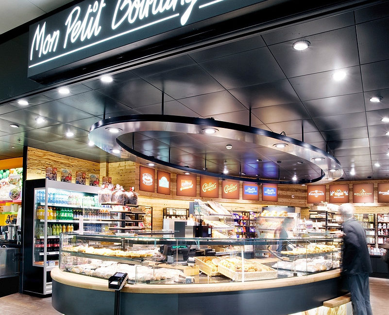 Un comptoir de boulangerie moderne portant l'enseigne « Mon Petit Boulanger Chéri », fabriqué en bois massif, présente divers viennoiseries et pains, tandis qu'une personne fait son choix et que, en arrière-plan, on aperçoit des rayons de boissons et d'autres produits. - Menuiserie Ledermann SA