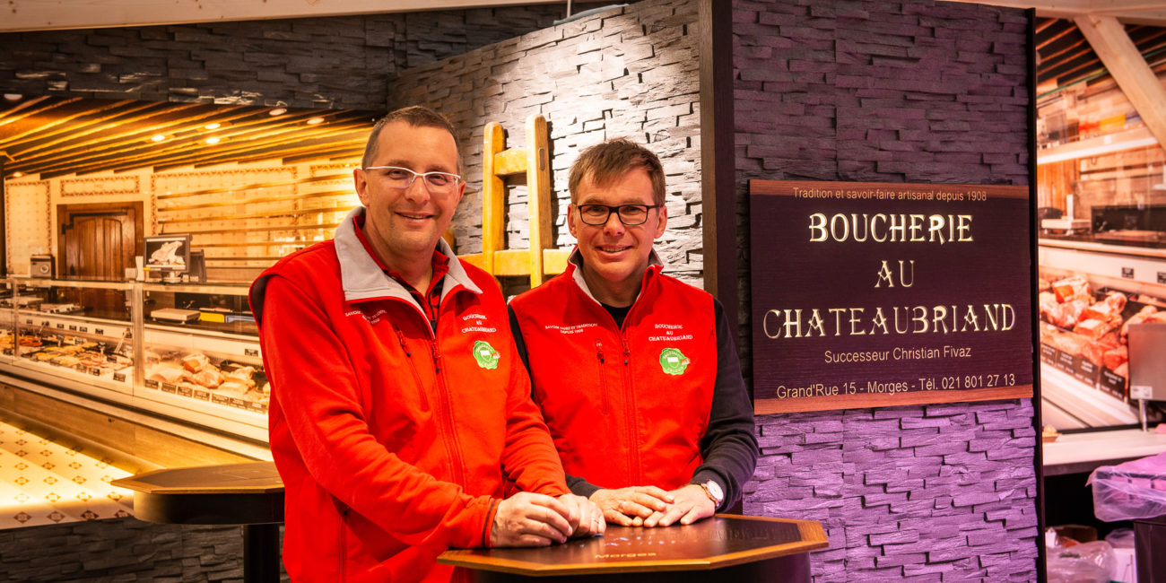 Deux hommes vêtus de vestes rouges se tiennent côte à côte, souriants, à l'intérieur de la « Boucherie au Chateaubriand ». L'agencement intérieur du magasin se caractérise par d'élégantes vitrines et met en valeur, en arrière-plan, des éléments de menuiserie sur mesure. - Menuiserie Ledermann SA