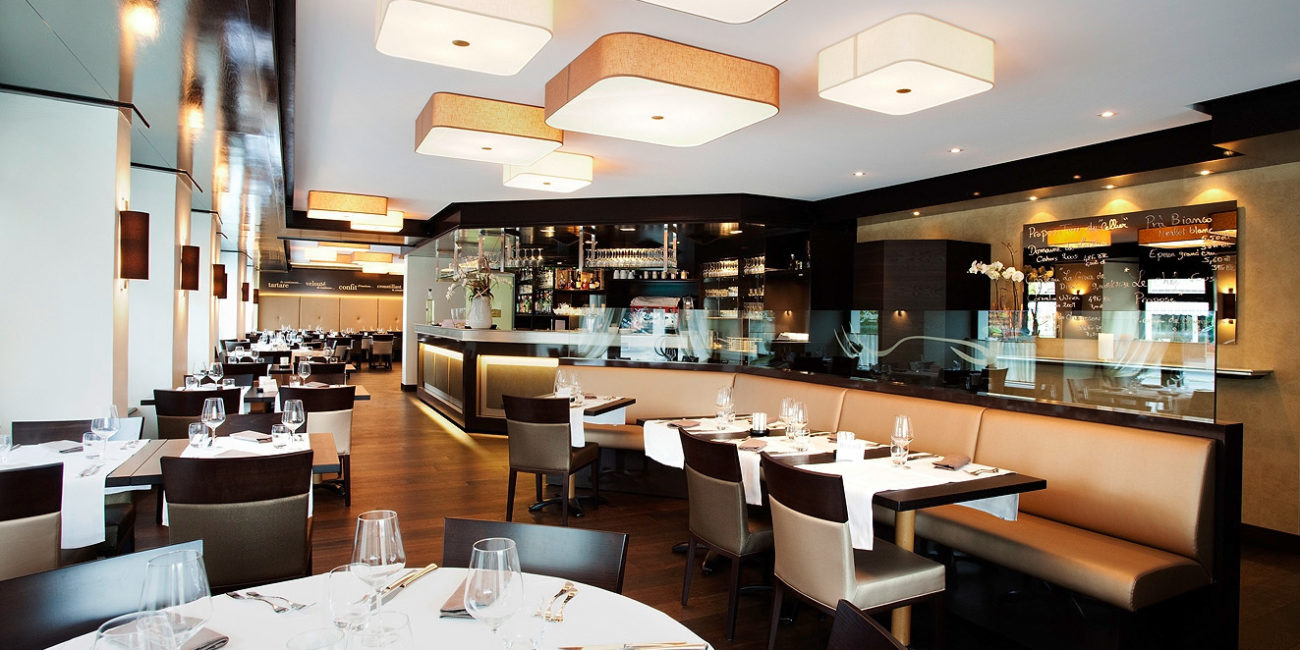 Intérieur de restaurant moderne avec des tables soigneusement dressées, des chaises beiges et des verres à vin. Des suspensions géométriques et une cloison en verre rehaussent l'ambiance, tandis que des touches de menuiserie sur mesure subliment l'agencement intérieur. - Menuiserie Ledermann SA