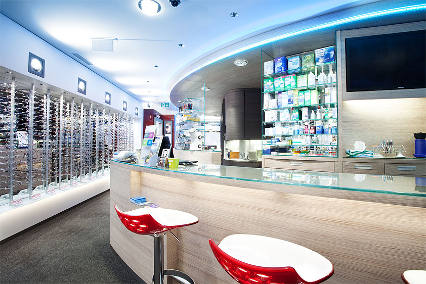 Intérieur moderne d'un magasin d'optique, avec un comptoir en verre, deux tabourets rouges et blancs, des lunettes exposées sur le mur de gauche et, derrière le comptoir, des étagères en bois massif remplies de lunettes et d'accessoires. - Menuiserie Ledermann SA