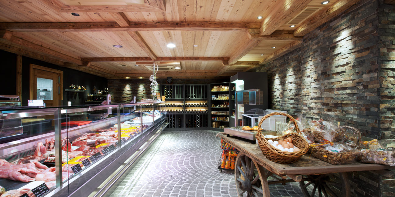 Une boucherie moderne au sol en pierre et au plafond en bois massif, dont l'aménagement intérieur comprend une vitrine à viande en verre sur la gauche et un chariot rustique en bois chargé de paniers d'œufs ; des étagères et des bouteilles de vin occupent l'arrière-plan. - Menuiserie Ledermann SA