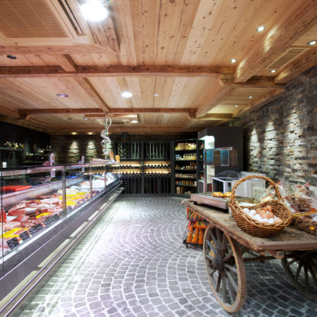 Une boucherie moderne au sol en pierre et au plafond en bois massif, dont l'aménagement intérieur comprend une vitrine à viande en verre sur la gauche et un chariot rustique en bois chargé de paniers d'œufs ; des étagères et des bouteilles de vin occupent l'arrière-plan. - Menuiserie Ledermann SA