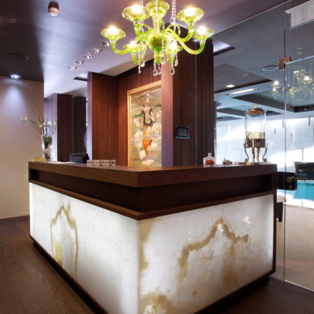 Un comptoir d'accueil orné d'une façade en onyx lumineuse trône dans une pièce moderne et tamisée. Des éléments de menuiserie sur mesure et des touches de bois massif mettent l'espace en valeur, tandis qu'un lustre vert, un tableau, une fontaine à eau en verre et un vase orné de branches apportent une touche d'élégance. - Menuiserie Ledermann SA