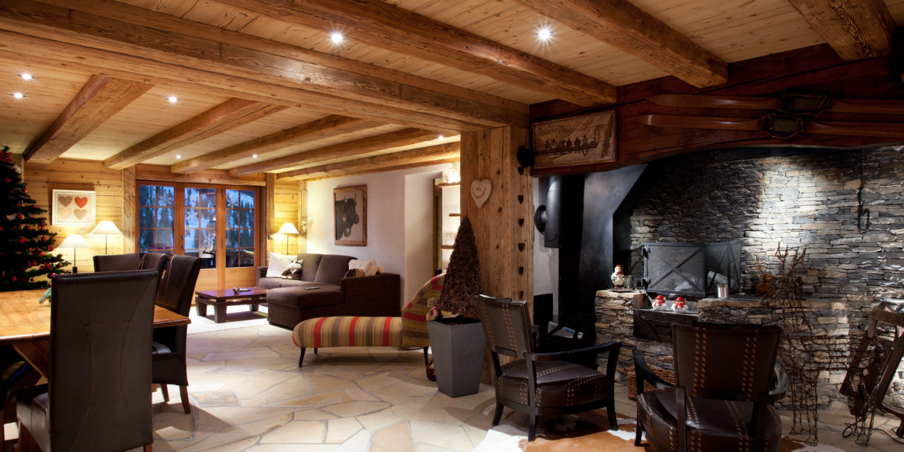 Un salon chaleureux avec des poutres en bois massif, une cheminée en pierre, des fauteuils en cuir, un sapin de Noël décoré, de grandes fenêtres et une table à manger — le tout magnifiquement mis en valeur par un éclairage chaleureux et des éléments de menuiserie sur mesure. - Menuiserie Ledermann SA