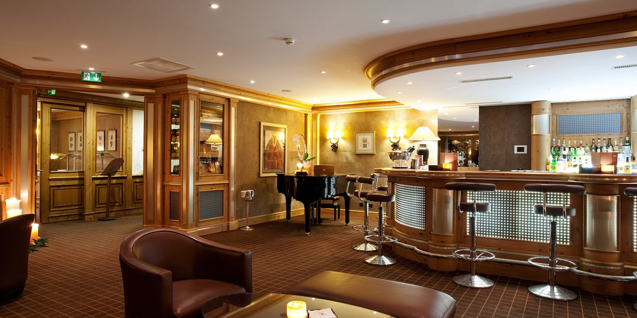 Un bar d'hôtel doté d'un comptoir incurvé en bois massif, de tabourets de bar modernes, d'un piano à queue, de fauteuils en cuir et de tables éclairées à la lueur des bougies. Un éclairage chaleureux, des boiseries inspirées de la menuiserie sur mesure et des œuvres d'art encadrées agrémentent l'espace. - Menuiserie Ledermann SA