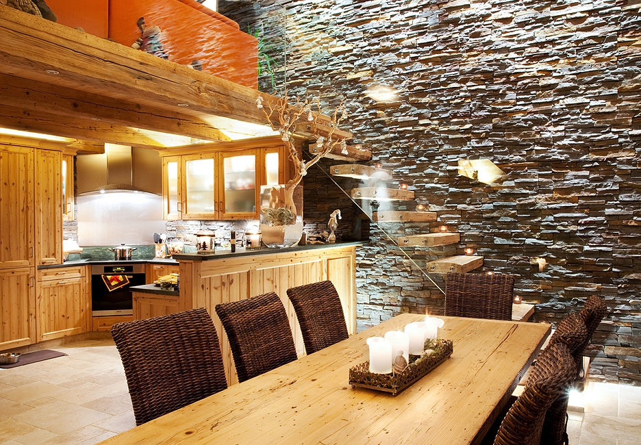 Une salle à manger au style rustique moderne, avec une longue table en bois massif et des chaises en osier, ouverte sur une cuisine dotée d'armoires sur mesure en menuiserie et d'un mur d'accent en pierre. Un escalier vitré mène à une mezzanine, tandis que des plafonniers diffusent une lumière chaleureuse. - Menuiserie Ledermann SA