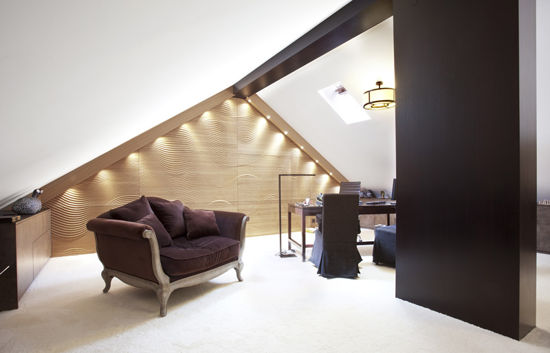 Cette chambre mansardée moderne est aménagée avec une causeuse marron, un bureau en bois massif accompagné d'une chaise, des panneaux muraux à la texture ondulée, un éclairage encastré, un plafond en pente doté d'une lucarne et une moquette beige, pour un agencement intérieur élégant. - Menuiserie Ledermann SA