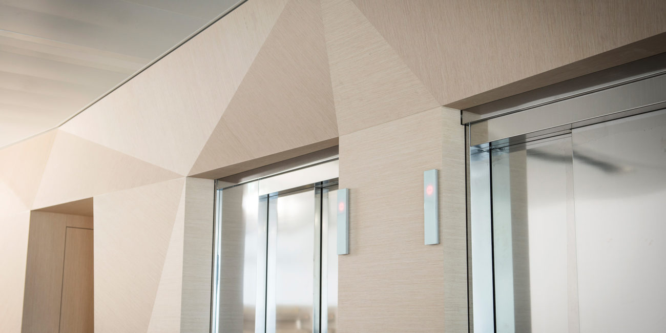 Deux portes d'ascenseur modernes aux finitions métalliques s'intègrent dans un mur beige orné de motifs géométriques et angulaires, rehaussé par des touches de menuiserie sur mesure. Deux boutons d'appel d'ascenseur lumineux sont fixés au mur entre les deux portes. - Menuiserie Ledermann SA