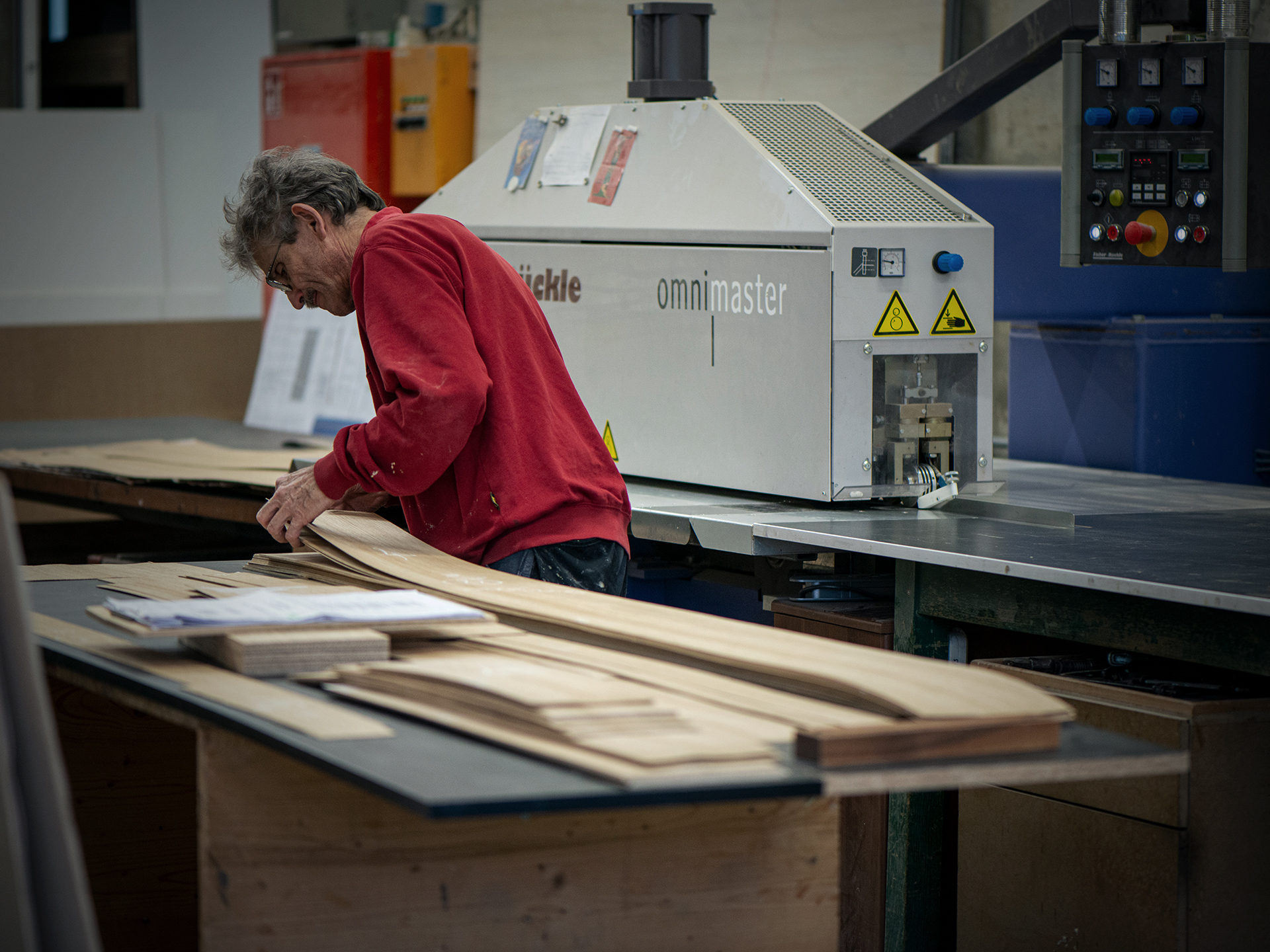 Une personne vêtue d'un pull rouge examine et dispose de fines planches en bois massif sur un établi, dans un atelier où s'alignent des machines industrielles et des outils. - Menuiserie Ledermann SA