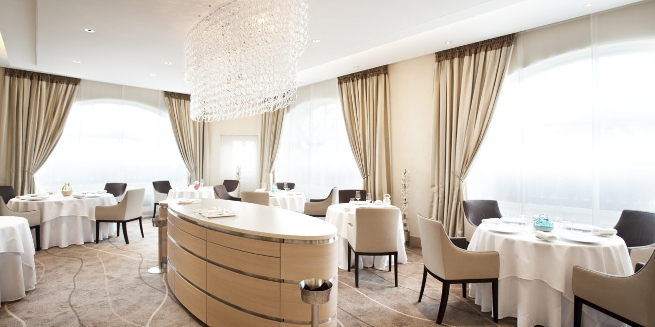 Une salle de restaurant moderne et lumineuse, avec des tables rondes recouvertes de nappes blanches, des chaises rembourrées beiges, de grandes fenêtres ornées de rideaux et un comptoir central en bois massif, sous un lustre en cristal. - Menuiserie Ledermann SA