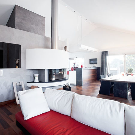 Un salon-salle à manger moderne agrémenté d'un canapé blanc et rouge, d'une cheminée cylindrique, d'un parquet en bois massif, d'une grande table à manger avec des chaises noires, ainsi que de baies vitrées ornées de voilages, pour un agencement intérieur raffiné. - Menuiserie Ledermann SA