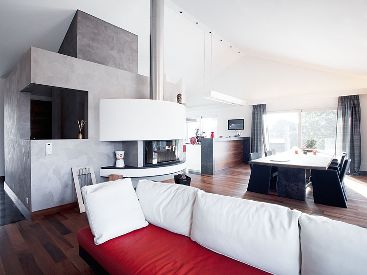 Un salon-salle à manger moderne agrémenté d'un canapé blanc et rouge, d'une cheminée cylindrique, d'un parquet en bois massif, d'une grande table à manger avec des chaises noires, ainsi que de baies vitrées ornées de voilages, pour un agencement intérieur raffiné. - Menuiserie Ledermann SA
