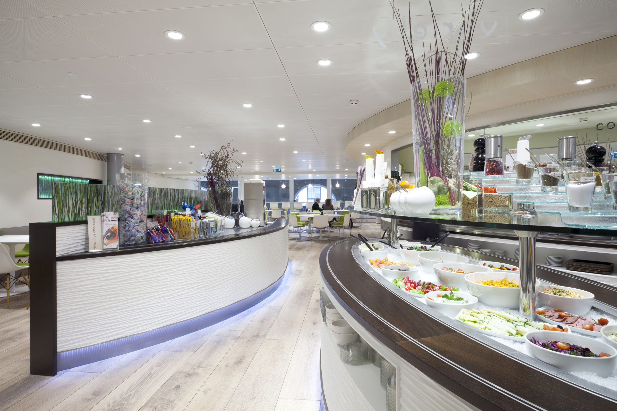 Entreprise 12 Un restaurant-buffet moderne doté d'un comptoir de service incurvé, de saladiers et de garnitures, de plantes décoratives, ainsi que de tables et de chaises en bois massif soigneusement disposées, le tout mis en valeur par un éclairage zénithal lumineux. - Menuiserie Ledermann SA