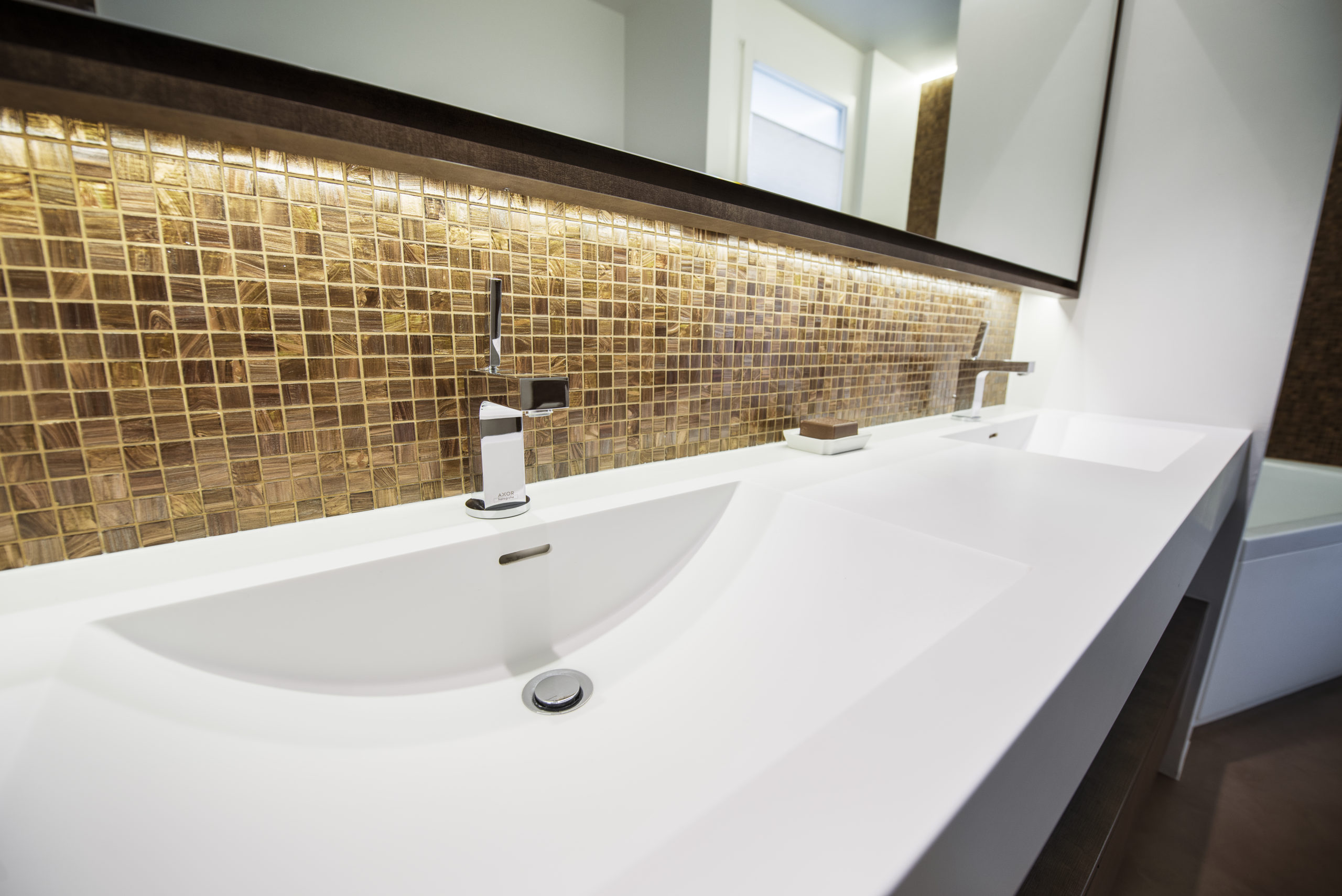 Entreprise 13 Un lavabo de salle de bains moderne, doté d'un plan de travail blanc, de deux robinets chromés, d'une crédence en mosaïque aux tons bruns et dorés, ainsi que d'un grand miroir avec éclairage intégré au-dessus, met parfaitement en valeur le savoir-faire de la menuiserie sur mesure. - Menuiserie Ledermann SA