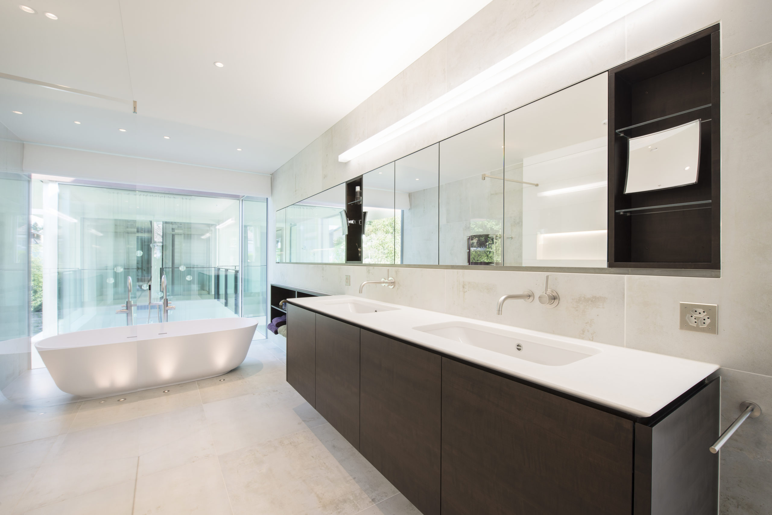 Entreprise 14 Une salle de bains moderne dotée d'un meuble-lavabo à double vasque en bois massif, de robinets muraux, de grands miroirs et d'une baignoire îlot située près de baies vitrées donnant sur la verdure. L'espace se caractérise par des tons neutres et un agencement intérieur minimaliste. - Menuiserie Ledermann SA