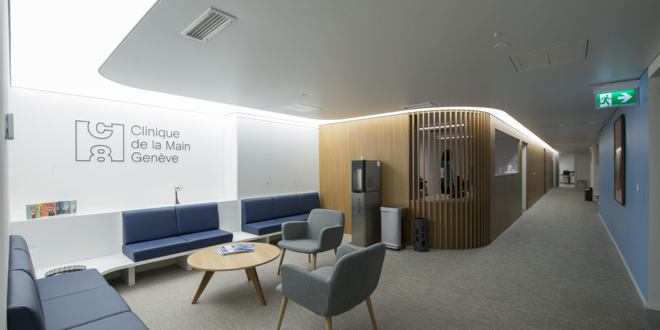 Une salle d'attente de clinique moderne, équipée de sièges bleus et gris, d'une table ronde en bois massif, d'un distributeur d'eau et d'une cloison. Le mur arbore le logo « Clinique de la Main Genève ». L'espace est bien éclairé, propre et met en valeur un aménagement intérieur haut de gamme. - Menuiserie Ledermann SA