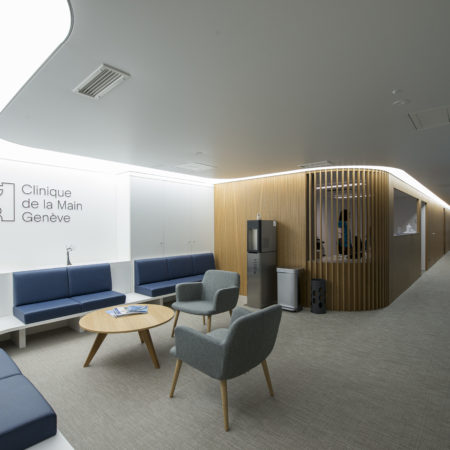 Une salle d'attente de clinique moderne, équipée de sièges bleus et gris, d'une table ronde en bois massif, d'un distributeur d'eau et d'une cloison. Le mur arbore le logo « Clinique de la Main Genève ». L'espace est bien éclairé, propre et met en valeur un aménagement intérieur haut de gamme. - Menuiserie Ledermann SA