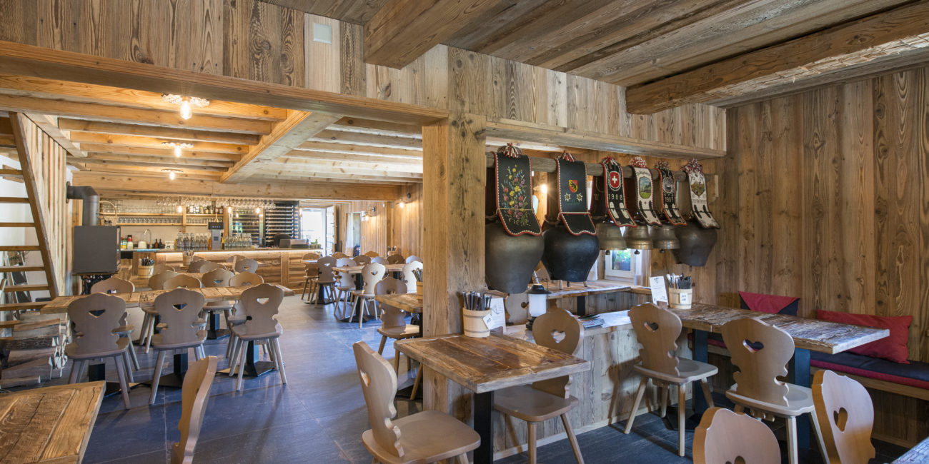 Intérieur spacieux d'un restaurant, avec des murs, un plafond et du mobilier en bois massif. Plusieurs tables et chaises en bois vides sont disposées un peu partout dans la salle. On aperçoit des éléments décoratifs et des pompes à bière sur la droite. - Menuiserie Ledermann SA