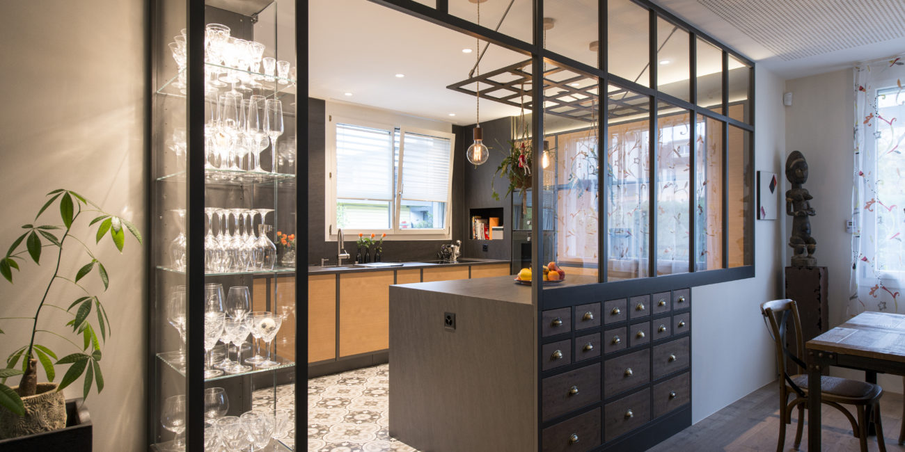 Une cuisine moderne avec un sol en carrelage à motifs, des vaisseliers en verre mettant en valeur la verrerie, des cloisons en verre à cadre noir, du bois massif et des meubles en bois clair, ainsi qu'un coin repas avec une table et des chaises en bois. - Menuiserie Ledermann SA