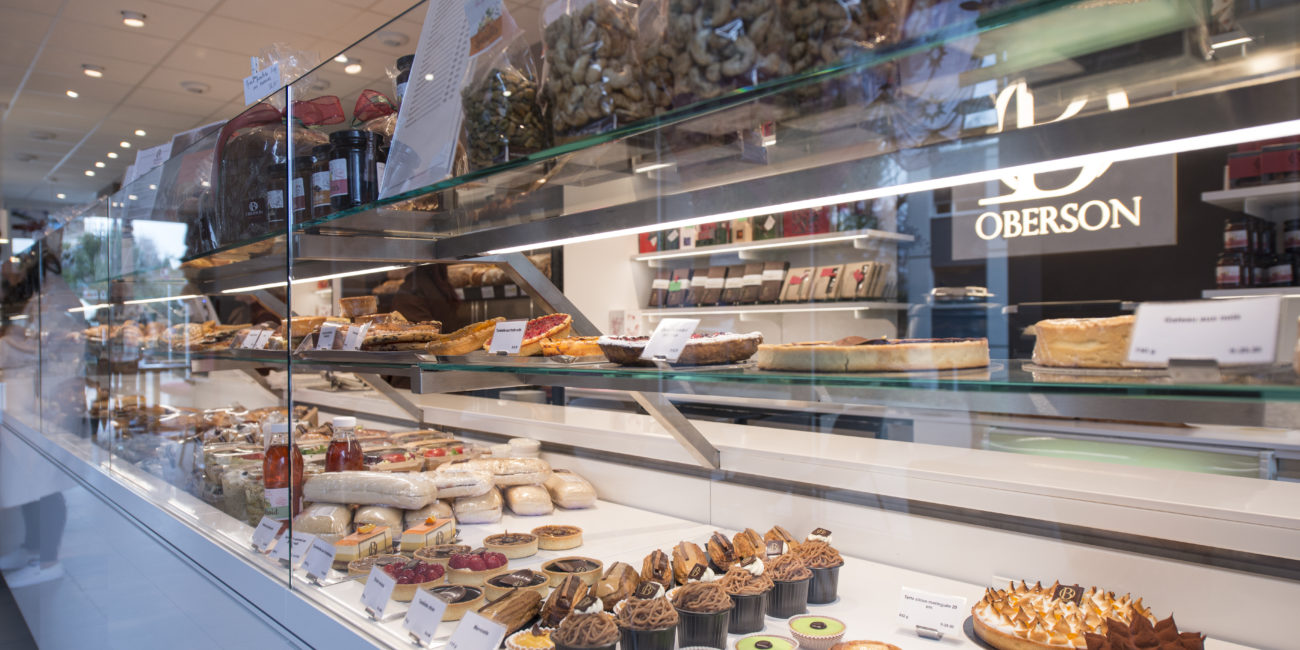 Dans une boulangerie ou une pâtisserie, une vitrine en verre met en valeur les gâteaux, les tartes et les pâtisseries disposés sur des étagères sur mesure en bois massif, alliant un agencement intérieur élégant à de délicieuses pâtisseries et à des produits soigneusement étiquetés en arrière-plan. - Menuiserie Ledermann SA